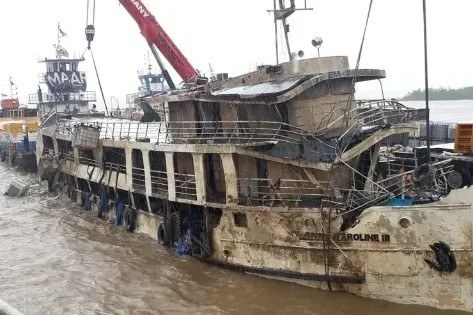 A imagem registra a complexa operação de resgate do navio Anna Karoline III em águas turvas no Amapá, exibindo a embarcação parcialmente submersa e com visíveis danos estruturais em seus múltiplos conveses. O casco e as grades de proteção apresentam-se cobertos de lama e detritos, enquanto um guindaste vermelho de grande porte domina a parte superior da cena, utilizando cabos de aço para içar ou estabilizar a estrutura comprometida. Ao fundo, sob um céu nublado, outras embarcações de apoio participam da logística, evidenciando o esforço técnico e a gravidade do naufrágio em meio ao cenário fluvial.