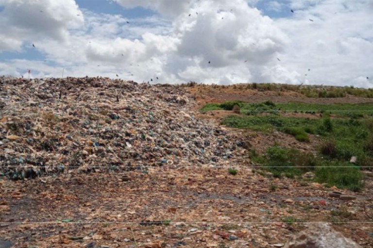 Uma montanha de resíduos descartados a céu aberto em um lixão, com urubus sobrevoando a área sob um céu com nuvens carregadas e vegetação rasteira ao lado da pilha de lixo.