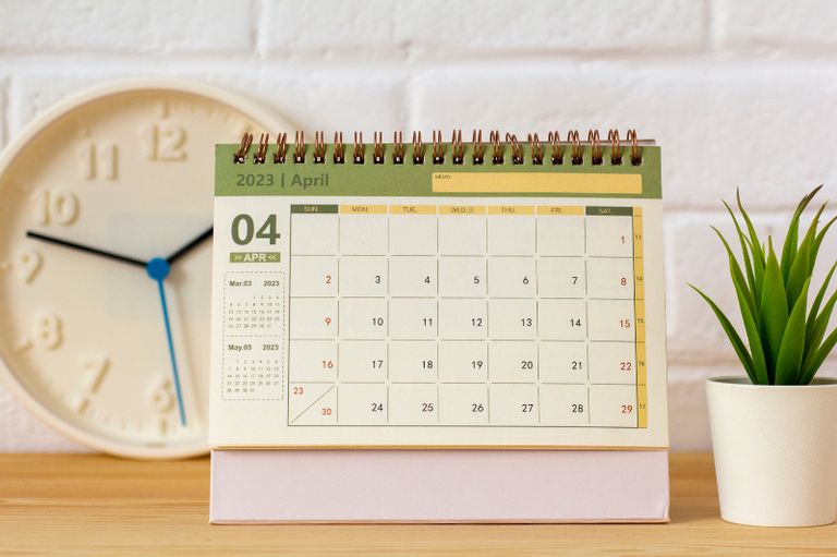 Um relógio branco, um calendário de abril marcando o dia 4, e uma pequena planta em um vaso branco sobre uma mesa de madeira, em frente a uma parede de tijolos brancos.