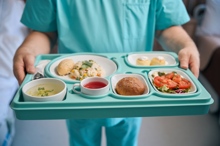 Foto de uma pessoa em uniforme hospitalar segurando uma bandeja verde-água com uma refeição completa, incluindo sopa, arroz, salada e sobremesa.