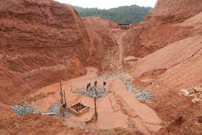 A imagem anexa mostra uma vista panorâmica de uma área de mineração ou garimpo a céu aberto, caracterizada por uma profunda escavação no solo de cor vermelha ou ocre. As paredes íngremes e elevadas da cava revelam o intenso impacto ambiental da atividade. No centro da parte inferior, alguns trabalhadores (três ou quatro) estão envolvidos em atividades em torno de uma estrutura rudimentar de madeira, possivelmente para processamento ou extração de minério (como ouro), em meio à lama vermelha e cascalho. Um rastro de material e água escorre pelo centro do barranco, descendo para onde os garimpeiros estão. No topo da formação, ao longe, é possível ver um pedaço de vegetação e o que parece ser maquinário pesado, como um trator ou escavadeira, indicando a continuidade da operação. O ambiente geral é de grande perturbação da paisagem natural.