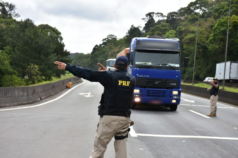 Policial rodoviário federal, de costas, faz sinal de parada para um caminhão azul em uma rodovia cercada por vegetação. Ao fundo, um segundo agente auxilia na orientação do trânsito.