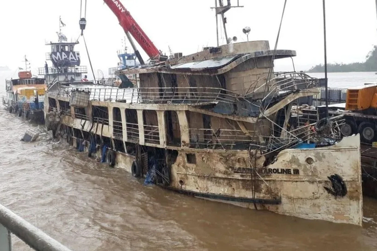 Embarcação identificada como Anna Caroline III, parcialmente submersa em águas barrentas. Um guindaste vermelho está sendo usado para tentar içar a embarcação destruída, que se encontra ao lado de outras duas embarcações.
