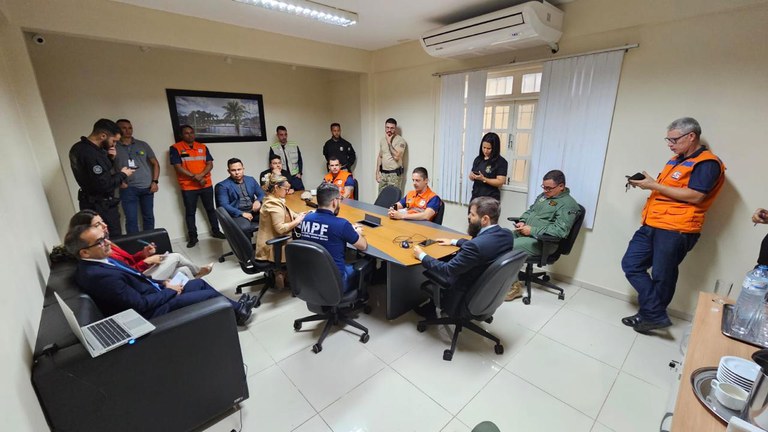 Dezoito pessoas, entre homens e mulheres, estão em uma sala de reuniões. Sete estão sentadas em cadeiras, ao redor de uma mesa, duas estão em um sofá e nove estão de pé