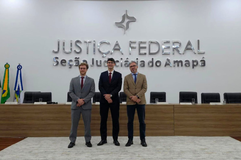 Três homens de terno e gravata posam sorrindo lado a lado no centro de um plenário. Ao fundo, uma parede branca exibe o letreiro metálico "JUSTIÇA FEDERAL - Seção Judiciária do Amapá" abaixo de um logotipo.