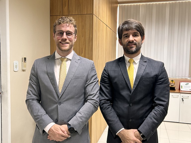 Dois homens sorriem para a foto. Eles estão de pé e usam terno