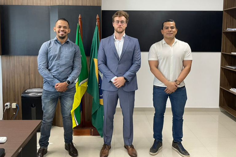 Três homens em pé, alinhados em uma sala de escritório, vestindo trajes sociais e casuais. Ao centro, entre eles, estão mastros com as bandeiras do Brasil e do Amapá.