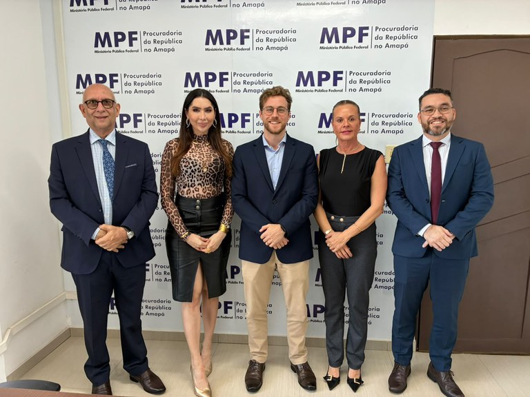 Cinco pessoas — três homens e duas mulheres em trajes formais — posam alinhadas e sorrindo para a foto.
Ao fundo, uma parede branca exibe um painel com logomarcas repetidas da Procuradoria da República no Amapá (MPF).