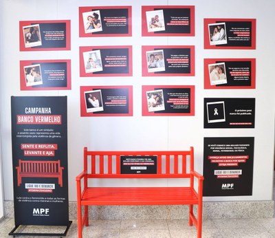 Registro da campanha Banco Vermelho instalada no hall da sede do MPF/BA. Ao centro, há um banco pintado de vermelho, acompanhado de um totem que explica seu significado como símbolo de enfrentamento à violência contra a mulher. Ao redor, estão expostos relatos fictícios sobre diferentes formas de violência contra a mulher. À direita, há um painel com orientações para oferecer apoio a mulheres em situação de violência e informações sobre o Ligue 180.