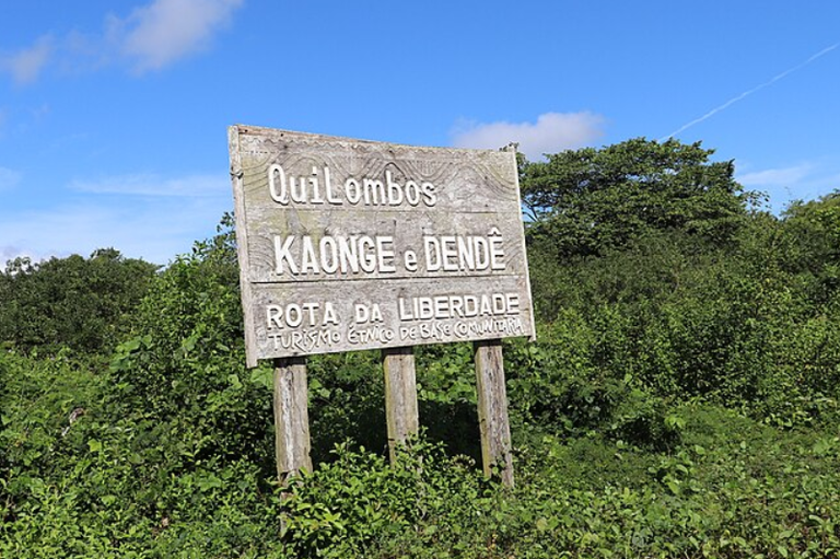 Foto mostra placa de madeira em meio à vegetação com os dizeres "Quilombos Kaonge e Dendê - Rota da Liberdade - Turismo Étnico de Base Comunitária", em dia claro de sol e céu azul.