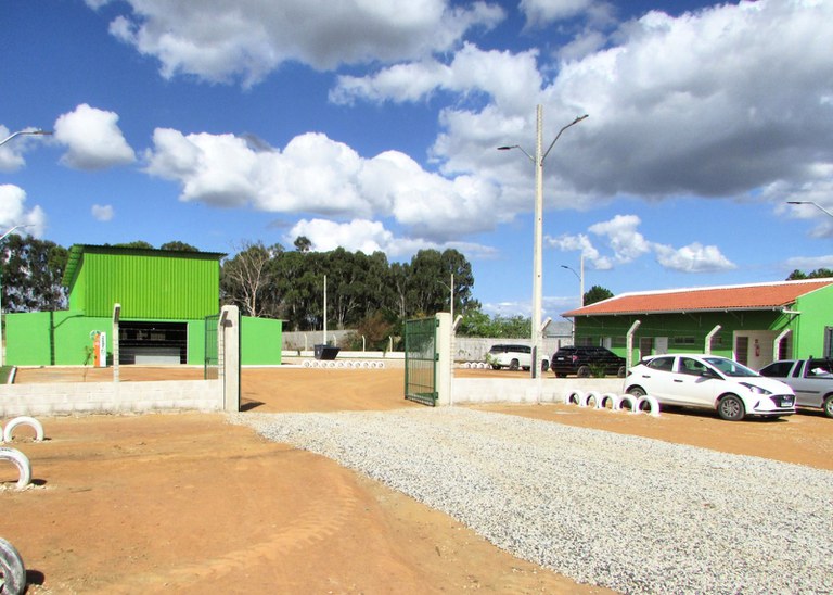 Foto com vista geral de um centro de reciclagem com galpão verde, área de cascalho e estacionamento delimitado por pneus brancos. O espaço é aberto, murado e tem carros estacionados.