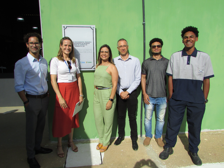 Grupo de seis pessoas, composto por quatro homens e duas mulheres. Todos estão em pé, vestindo roupas formais e sorrindo enquanto olham para frente. Atrás do grupo há uma placa de inauguração presa a uma parede na cor verde. Todo o espaço é iluminado a luz ambiente.