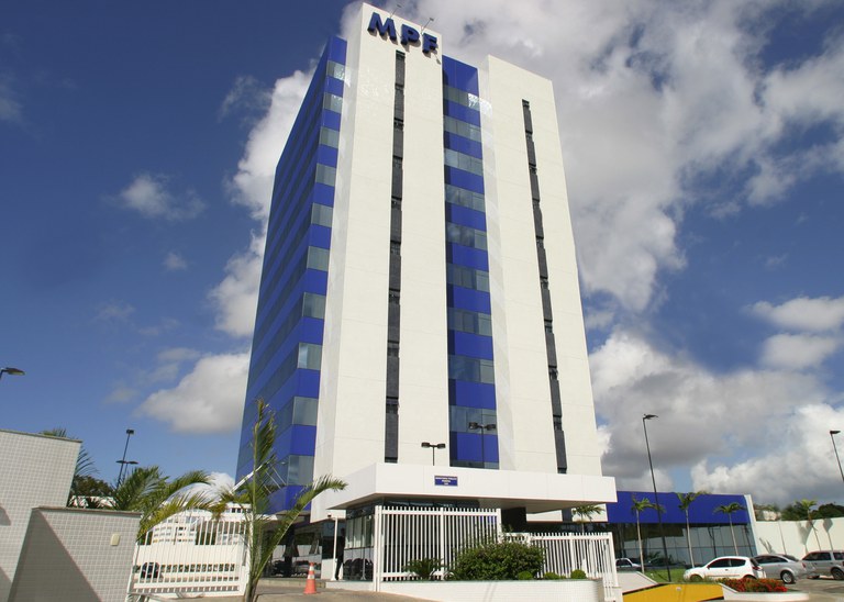 Fotografia diurna em plano inteiro mostrando a fachada de um prédio comercial alto e moderno com a sigla "MPF" no topo, sob um céu azul ensolarado com poucas nuvens.
