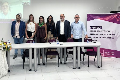 Na imagem há um grupo de três homens e três mulheres em pé, lado a lado dentro de uma sala branca. A frente deles há uma mesa com cadeiras e do lado direito um banner nas cores branco e roxo.