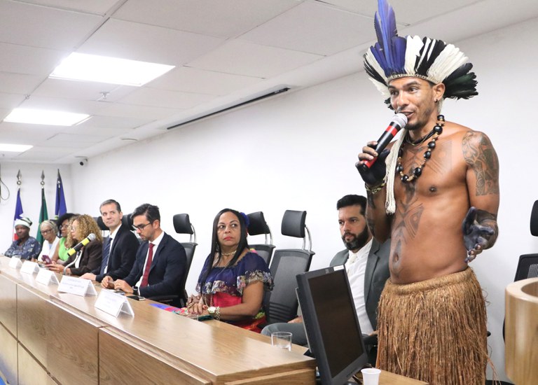 No auditório do MPF, um homem indígena com cocar de penas azuis, pretas e brancas, colar e saia de palha discursa ao microfone em pé, à frente de uma mesa composta por oito pessoas. Os demais participantes estão sentados atrás de uma bancada de madeira, com placas de identificação. Ao fundo, há bandeiras do Brasil e da Bahia.