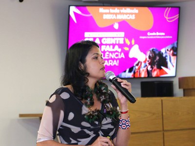 MPF Camilla Batista, secretária de Políticas para as Mulheres do Estado da Bahia, fala ao público durante a palestra que antecedeu a inauguração da campanha Banco Vermelho. Ela segura um microfone e está diante de uma tela que exibe a apresentação “Nem toda violência deixa marcas”. Ao fundo, aparecem elementos da identidade visual da palestra e uma imagem relacionada ao tema da violência contra a mulher.