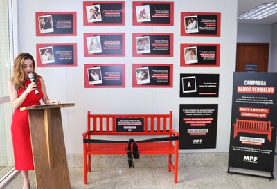 A procuradora da República Melina Montoya faz seu discurso durante a solenidade de inauguração da campanha Banco Vermelho. Ela está ao lado de um púlpito de madeira, segurando um microfone, em frente à instalação da campanha no hall da sede do MPF/BA. Ao centro, aparece o Banco Vermelho, cercado por uma faixa preta durante a solenidade. Ao fundo, estão expostos os relatos da campanha.