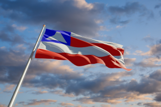Bandeira do estado da Bahia tremulando ao vento em um mastro, com o céu ao fundo repleto de nuvens em tons de azul, cinza e dourado. A bandeira apresenta listras horizontais vermelhas e brancas, além de um retângulo azul no canto superior esquerdo com um triângulo branco central.