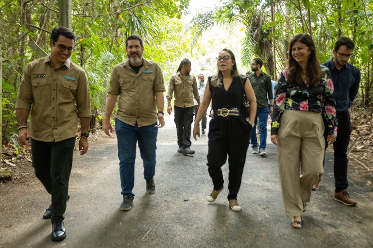 Grupo de pessoas caminha por trilha cercada de vegetação. Alguns usam camisas do Inema, outros estão com roupas casuais. Todos seguem juntos em ambiente de visita técnica ao ar livre.