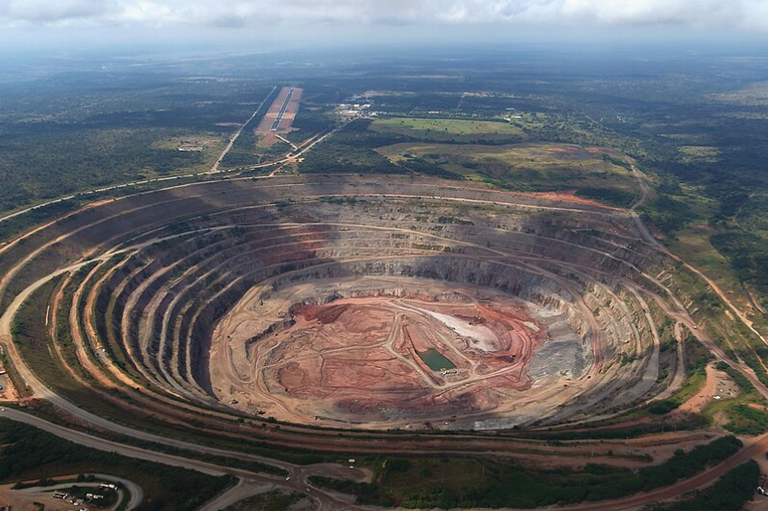 Vista aérea de uma gigantesca mina de diamantes a céu aberto, caracterizada por uma profunda cratera circular com múltiplas camadas de escavação em degraus. O fundo da mina apresenta solo em tons avermelhados e pequenos depósitos de água. Ao redor da mina, estende-se uma vasta paisagem de vegetação rasteira, com uma estrada e uma pista de pouso retilínea visíveis ao fundo, sob um céu com nuvens esparsas.