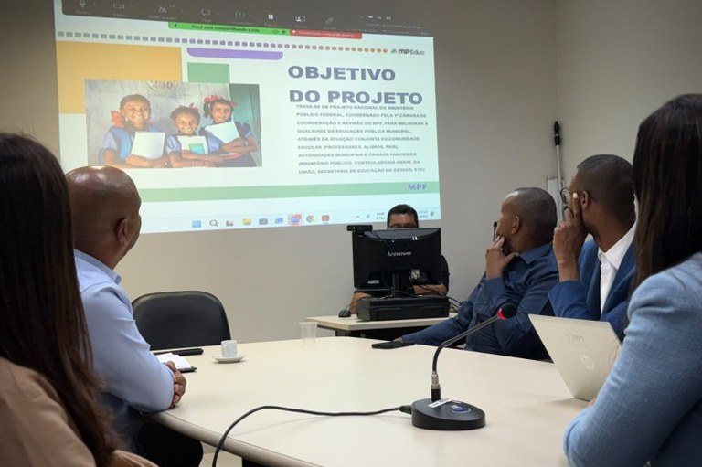 Foto mostra participantes da reunião em volta de uma mesa. Informações sobre o MPEduc são projetados em um telão