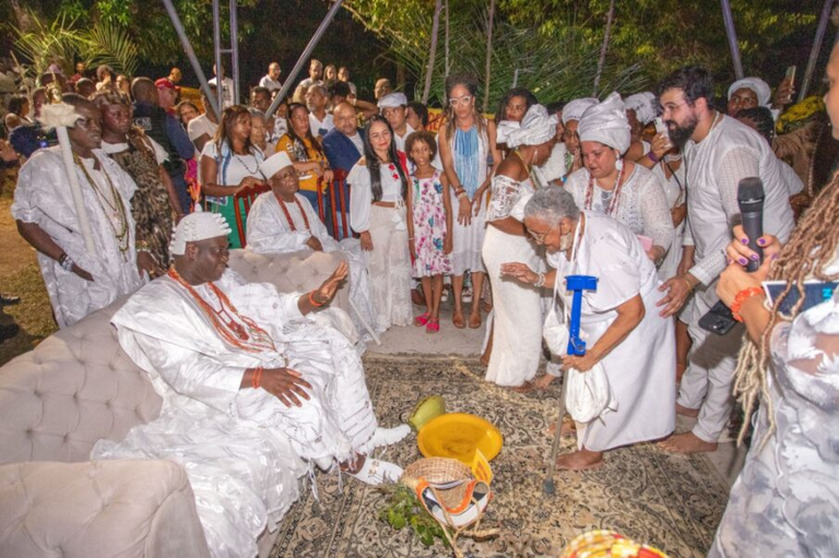 A imagem de cerimônia religiosa de matriz africana, com várias pessoas vestidas de branco. A atmosfera é de respeito e solenidade. Há elementos rituais no chão, como um recipiente amarelo e outros objetos. A cena se passa ao ar livre, à noite, com iluminação artificial.