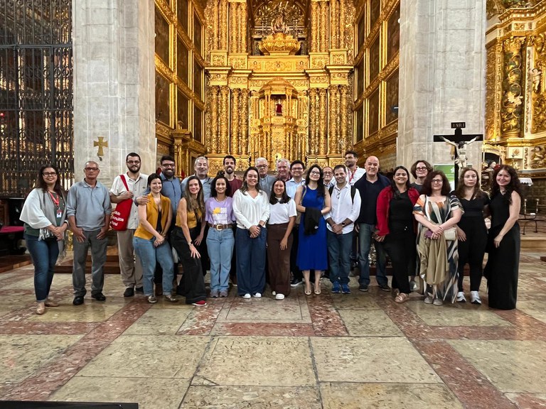 Um grupo de aproximadamente vinte e cinco pessoas posa reunido em frente a um altar ricamente ornamentado em ouro dentro de uma igreja histórica. A maioria está sorrindo, alinhada em uma fileira, algumas com roupas casuais e outras mais formais. O fundo é detalhado com colunas altas, esculturas religiosas e entalhes dourados que preenchem todo o altar. O chão é de mármore em tons claros e avermelhados. A atmosfera é iluminada e solene, destacando a arquitetura barroca do local.