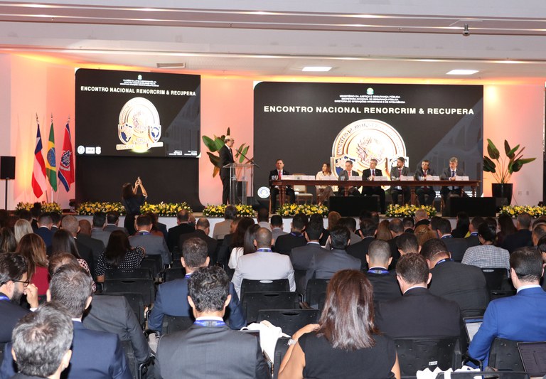 Imagem mostra um auditório cheio durante o “Encontro Nacional RENORCRIM & RECUPERA”, com palestrante no palco e autoridades sentadas à mesa acompanhando o evento.