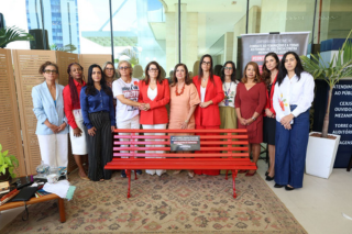 Grupo de mulheres em pé atrás de um banco vermelho com placa de campanha contra a violência às mulheres, em ambiente institucional, olhando para a câmera.