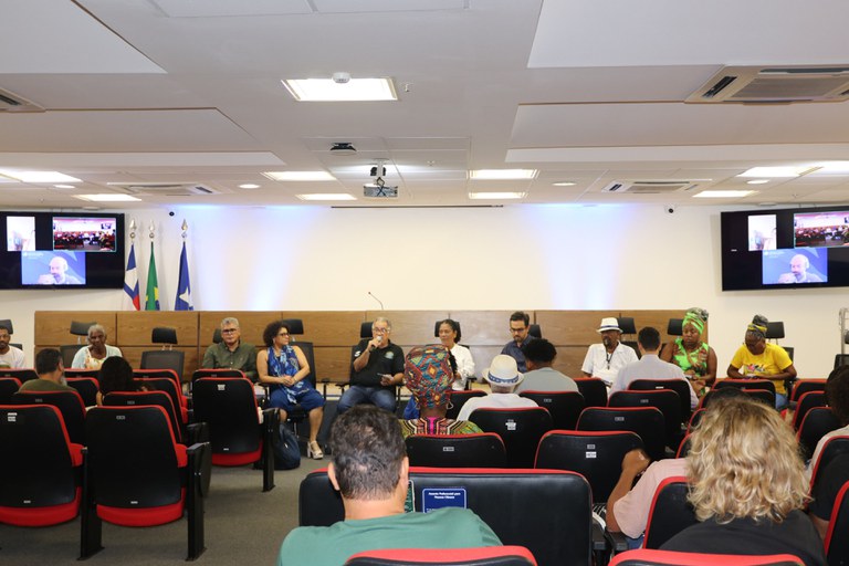 Esta imagem mostra uma apresentação ou painel de discussão ocorrendo em um auditório ou sala de conferências. Um grupo de oito pessoas está sentado em cadeiras de escritório atrás de uma longa mesa, de frente para a plateia. A plateia é composta por várias pessoas sentadas em fileiras de poltronas vermelhas, e a câmera está posicionada no fundo da sala, olhando para o palco. Há três monitores de tela plana visíveis: um projetor no centro (atualmente com uma tela branca) e dois monitores laterais que exibem a mesma imagem de um palestrante que não está fisicamente na sala. A sala é moderna, com tetos em painéis, ar-condicionado e luzes embutidas. As bandeiras do Brasil e de outros estados ou municípios estão visíveis no lado esquerdo do palco.