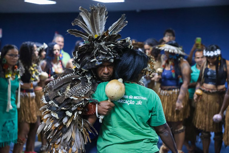 Foto de indígenas se abraçando. O que está de costas veste uma bluca verde onde está escrito Demarcação Já