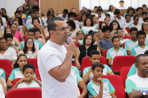 A imagem mostra um auditório cheio de pessoas, incluindo estudantes e adultos. No centro, um homem de pele morena, cabelos curtos e óculos está segurando um microfone e falando. Ele veste uma camiseta branca. As pessoas ao redor, sentadas em cadeiras vermelhas, têm expressões diversas, algumas prestando atenção, outras distraídas. Muitos estudantes estão com uniformes de camiseta branca e verde. O ambiente sugere um evento educacional ou uma palestra.