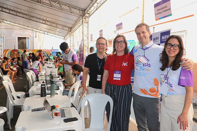 Foto tirada em evento do programa PopRuaJud, realizado em espaço aberto e coberto com tendas. À direita, quatro pessoas posam sorrindo para a câmera: duas mulheres (uma de camisa vermelha e outra com camisa do PopRuaJud), um homem de camisa preta com crachá e outro de camisa do PopRuaJud. Ao fundo, várias pessoas são atendidas em mesas com notebooks, copos e garrafas. Há placas identificando órgãos participantes, como o Ministério Público Federal (MPF) e a Defensoria Pública do Estado (DPE). O mural artístico ao fundo mostra rostos e elementos coloridos.