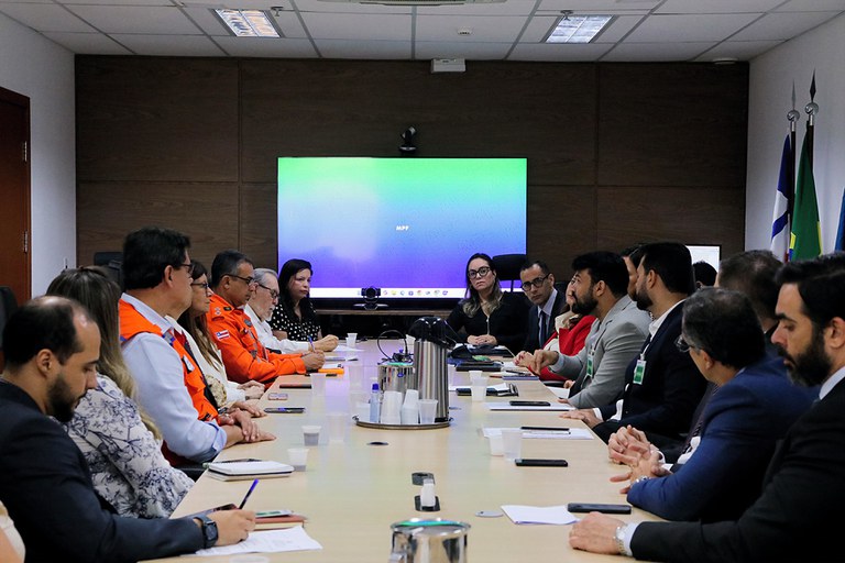 Foto de uma mesa de reunião com pessoas em volta e uma tela de TV ao fundo com um papel de parede azul, com a sigla MPF no centro
