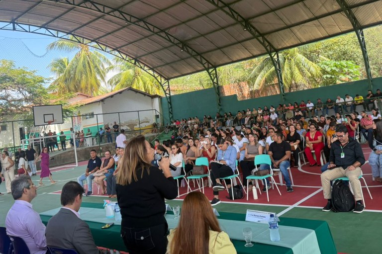 Uma mulher de cabelo loiro e blusa preta, de costas, fala ao microfone para uma plateia de dezenas de pessoas, sentadas em cadeiras azuis e brancas em uma quadra poliesportiva. Ao fundo da imagem, há coqueiros e um céu azul. Há também uma mesa retangular na parte inferior da imagem com algumas pessoas sentadas de frente em uma mesa de palestrantes.