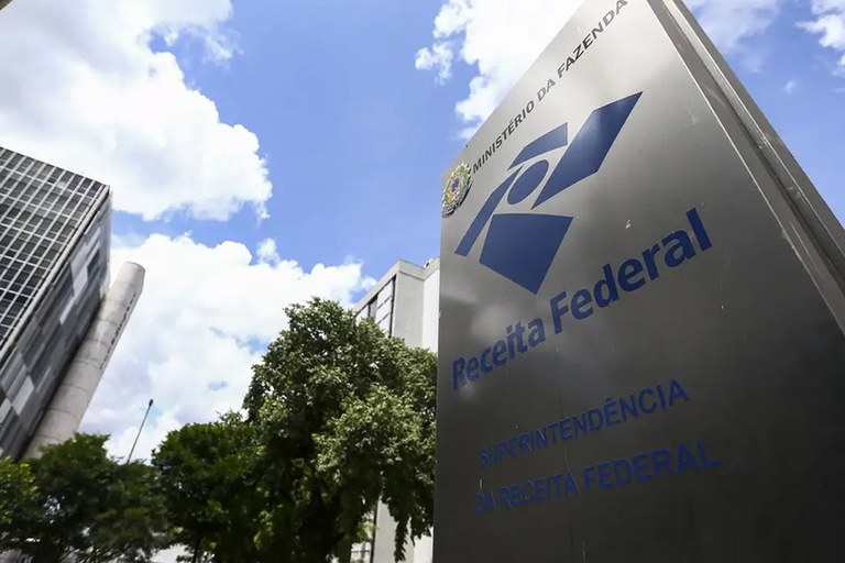 A imagem mostra a fachada de um prédio da Receita Federal do Brasil, com uma placa metálica em destaque contendo o logotipo do órgão e os dizeres "Ministério da Fazenda", "Receita Federal" e "Superintendência da Receita Federal". Ao fundo, aparecem edifícios, árvores e o céu com algumas nuvens.