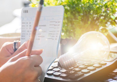 Foto da mão de uma pessoa que faz anotações numa caderneta. Atrás da caderneta, está um teclado de computador com uma lâmpada em cima.