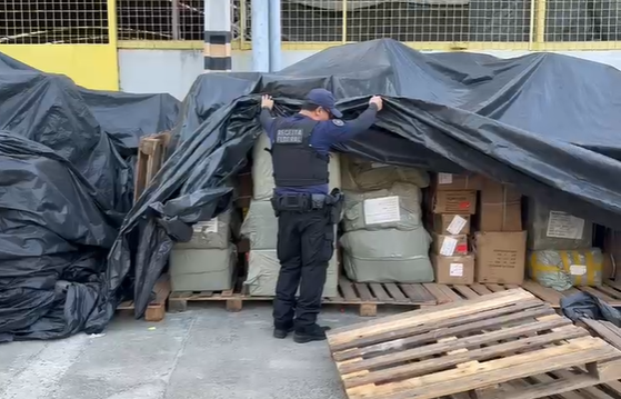 Um agente da Receita Federal, de costas, levanta uma lona preta que cobre grandes pilhas de caixas e pacotes. Ele veste uniforme azul-marinho e colete à prova de balas com a inscrição "RECEITA FEDERAL". Pallets de madeira estão espalhados pelo chão, e ao fundo, vê-se parte de um edifício.