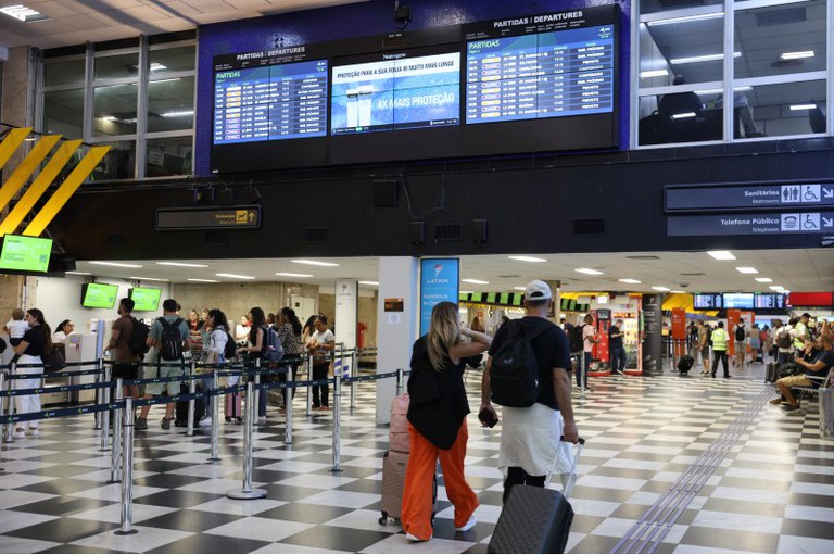 Imagem de passageiros em um aeroporto, alguns na fila do guichê da empresa aérea e outros passando
