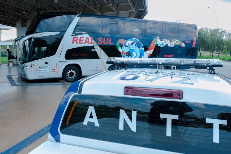 Viatura branca e azul da ANTT (Agência Nacional de Transportes Terrestres) em primeiro plano, com foco na parte traseira que exibe a sigla "ANTT" em letras brancas no vidro e a inscrição "FISCALIZAÇÃO FEDERAL" no teto próximo ao giroflex. Ao fundo, estacionado sob a estrutura coberta de um terminal, destaca-se um ônibus de viagem de dois andares da empresa "REAL SUL", com carroceria nas cores branca e azul escuro e estampa de um globo terrestre com bandeiras de países sul-americanos.