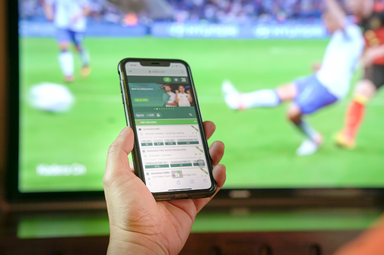 Uma mão segura um smartphone que exibe um aplicativo ou site de apostas esportivas. Ao fundo, uma televisão desfocada transmite uma partida de futebol em uma grande tela.
