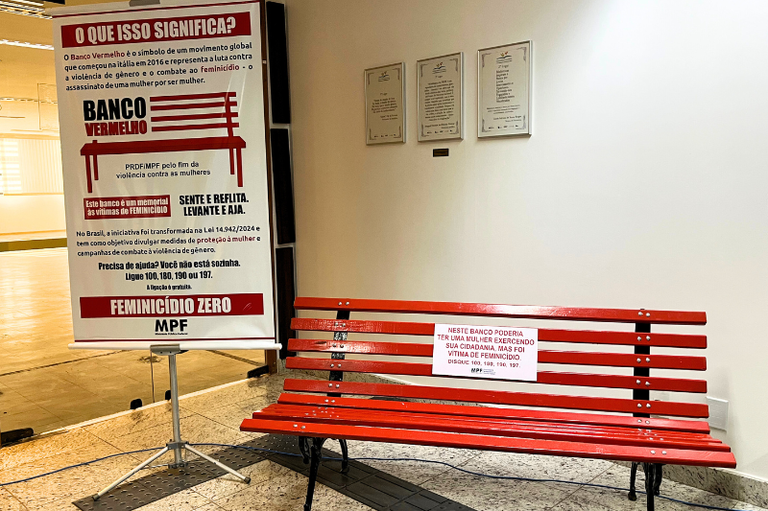 Imagem do banco vermelho de metal instalado na PR/DF. No banco há uma placa branca com a seguinte mensagem escrita em vermelho: “Neste banco poderia ter uma mulher exercendo sua cidadania, mas ela foi vítima de feminicídio”.
