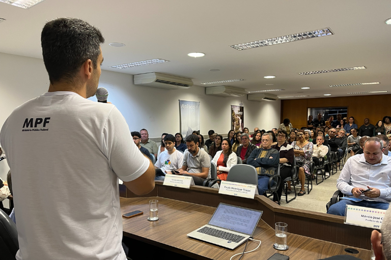Foto mostra um homem de costas com camiseta branca escrito MPF segurando um microfone enquanto fala com várias pessoas que estão a frente, sentadas, como em um auditório.
