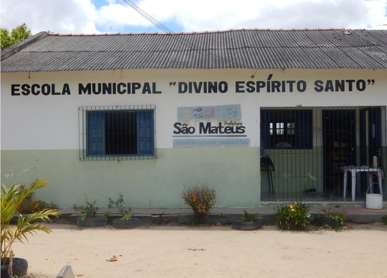 Fotografia mostra a fachada da escola municipal Divino Espírito Santo. A sede é uma casa simples, com grades na janela azul e na porta de entrada, além de telhado feito com telhas de amianto. É possível ver, em letras pretas pintadas na fachada, o nome da unidade de ensino e a logomarca da Prefeitura de São Mateus. A rua na frente da escola é de terra e há canteiros de flores plantados na frente.