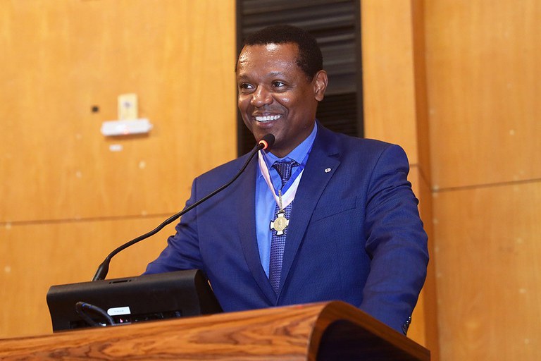 Foto de um homem negro vestindo terno escuro e uma medalha com as corees rosa e azul no peito. Ele fala ao microfone sobre um púlpito.