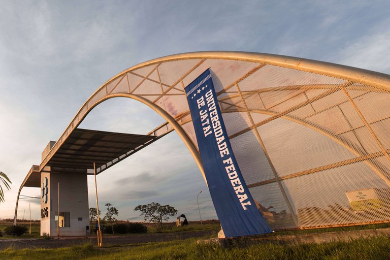 Fotografia do amanhecer do dia 21 de março de 2018 no pórtico de entrada da Universidade Federal de Jataí (GO). Há uma reua de acesso e uma estrutura metálica grande, em forma de arco, sobre uma guarita de alvenaria e uma faixa de pano azul no lado direito da estrutura com o testo "Universidade Federal de Jataí".