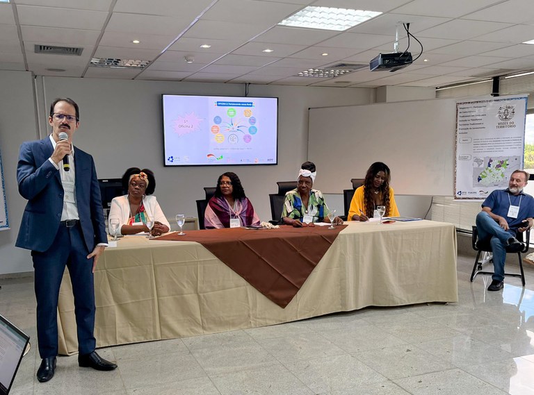 Fotografia de um evento institucional em uma sala de conferências. À esquerda, um homem branco de terno azul fala ao microfone. Atrás dele, quatro mulheres negras estão sentadas em uma mesa longa com toalha bege e detalhe marrom. Ao fundo, uma tela exibe um infográfico colorido e, à direita, há um banner com o título "Vozes do Território" e um mapa.