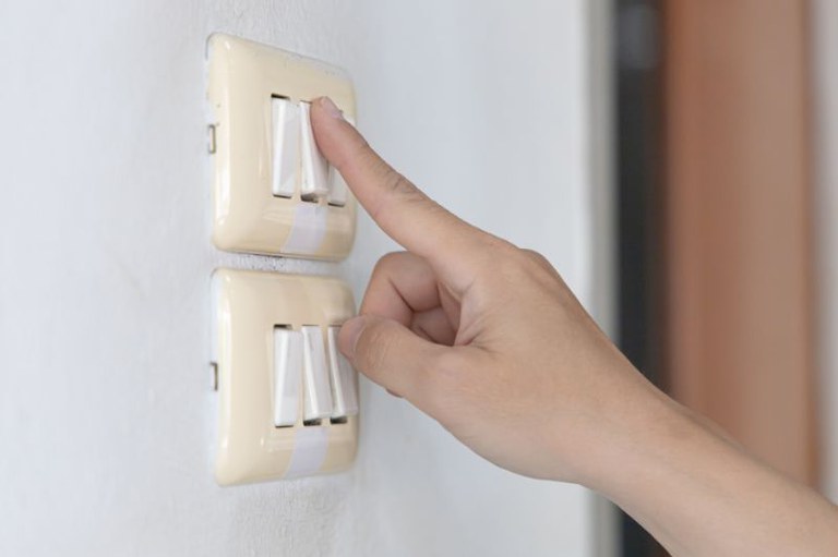 Foto mostra em detalhe uma pessoa acendendo um interruptor de tomada com três botões e abaixo outro interruptor com três botões
