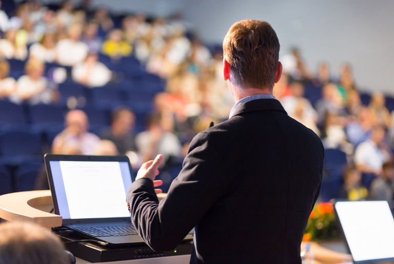 Uma pessoa está de pé em um pódio, fazendo uma apresentação para uma plateia em um grande auditório ou sala de conferências. A pessoa está de costas para a câmera, com um laptop aberto à sua frente exibindo um slide de apresentação. A audiência está sentada em fileiras de cadeiras, e a sala parece estar bem cheia, com muitas pessoas visíveis ao fundo.
