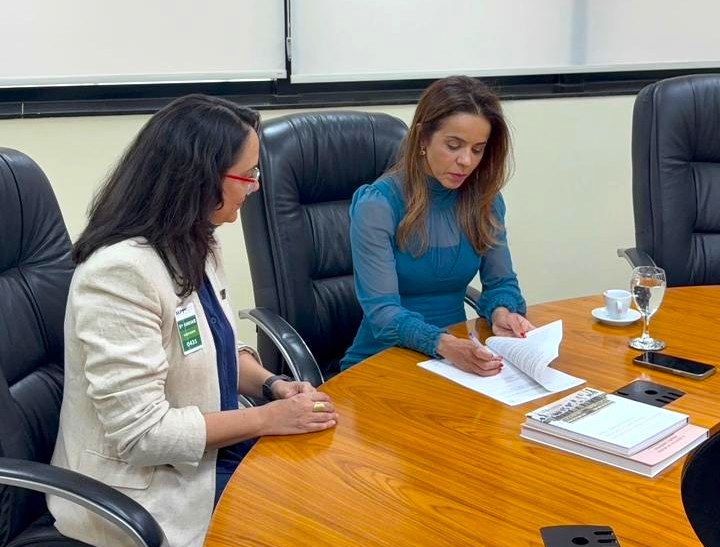 Duas mulheres estão sentadas a uma mesa oval de madeira em uma sala de reunião: a da direita, vestindo uma blusa azul-petróleo de mangas transparentes, está concentrada assinando um documento com caneta, enquanto a da esquerda, de blazer claro, óculos vermelhos e crachá de identificação, acompanha o ato atentamente. Sobre a mesa, encontram-se também uma pilha de livros, uma xícara de café, um copo d'água e um celular, tudo em um ambiente corporativo com cadeiras executivas pretas de couro ao fundo.